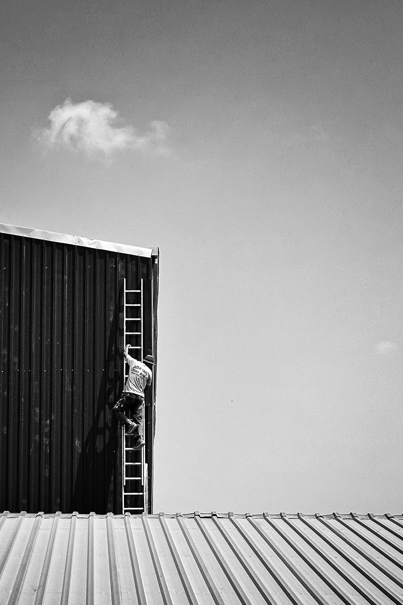 A worker ascends a vertical ladder against a corrugated building roof, monochrome shot.